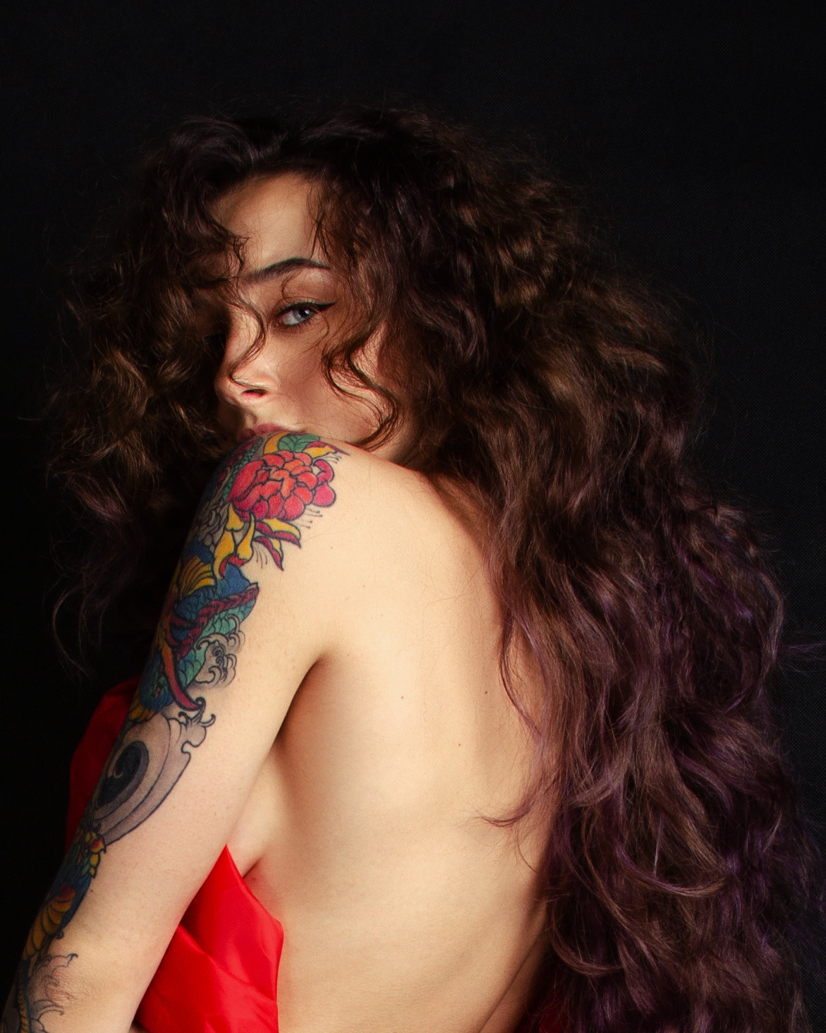 Портрет кудрявой девушки с открытой спиной в пол оборота к зрителю и с татуировкой на руке