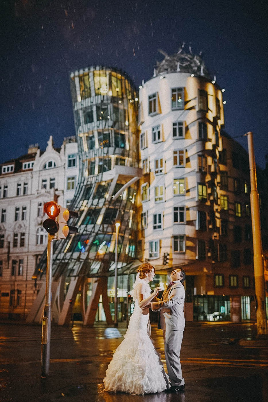 Same sex couple bride laughing as they embrace in the rain in front of the iconic Dancing House, or Ginger & Fred in Prague.