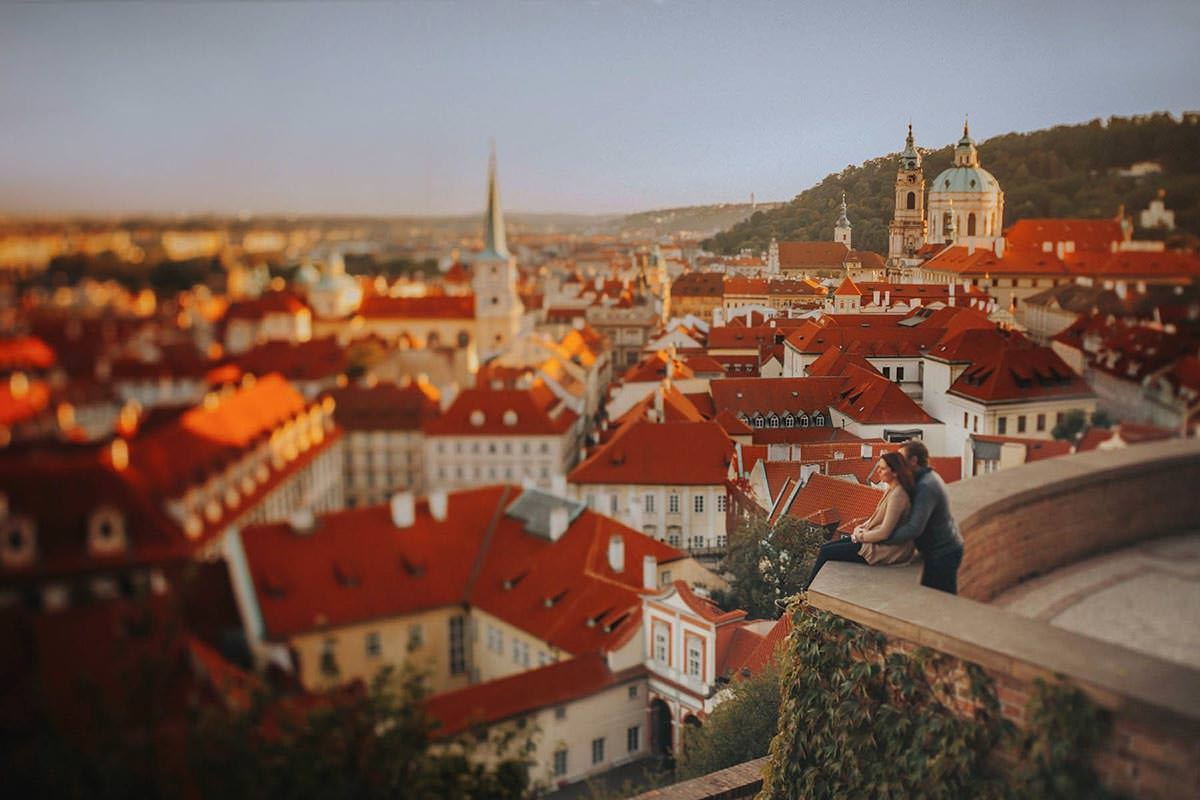 A tilt-shift lens distorts the skyline of Prague as the last rays of sunlight illuminate the red roofs as a couple sits above.