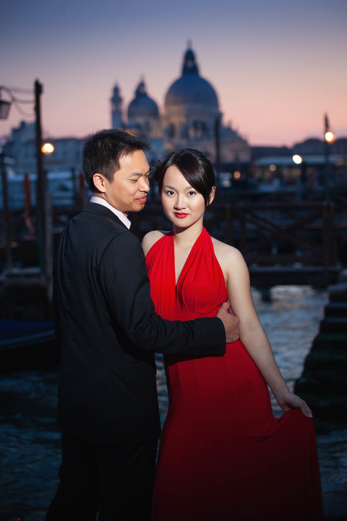 Thai bride in skin-tight red dress kissed by partner above gondola at sunset Venice.