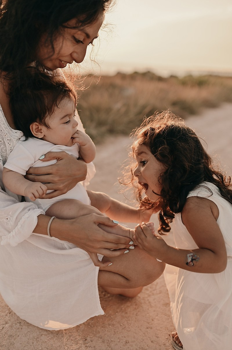 A mother holds her baby while the older daughter gently kisses the baby’s foot, all glowing in golden sunset light.