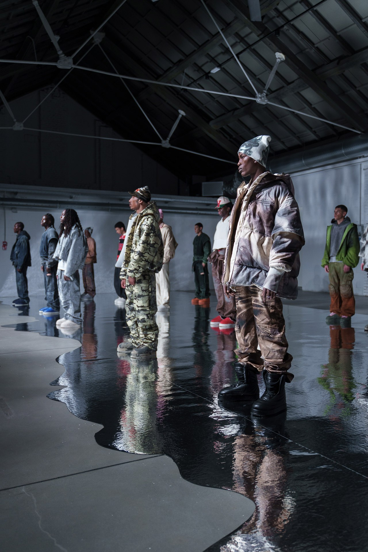A conceptual fashion presentation in an industrial setting. Models are lined up showcasing utilitarian streetwear, including camo prints and oversized silhouettes, reflected in a glossy, water-like floor installation.