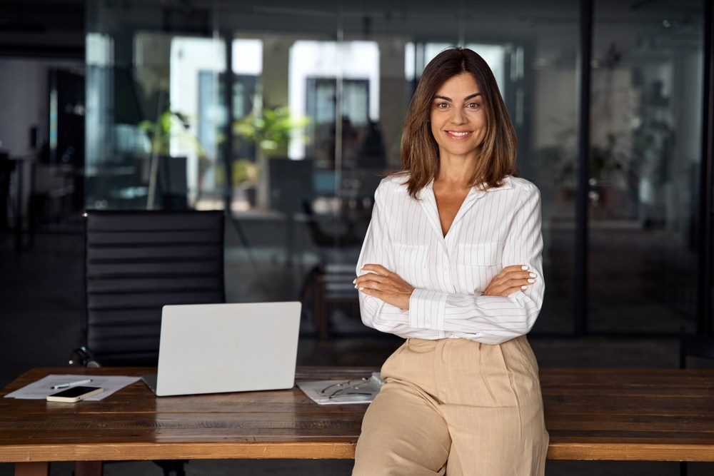 Smiling businesswoman leaning on a desk with arms crossed in a modern office environment