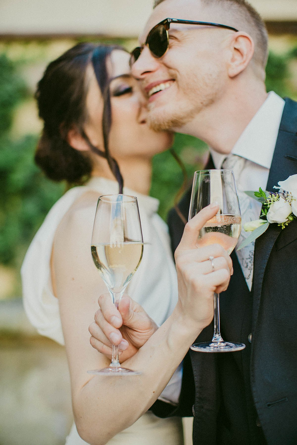 Joyful dark-haired bride kisses the cheek of her rayban sunglass wearing blonde groom while holding their champagne flutes in Prague.