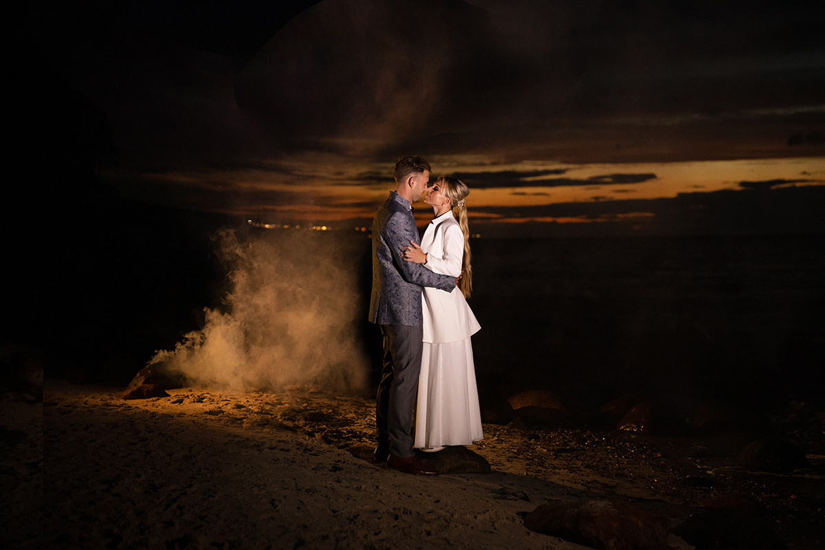 Das Brautpaar umarmt sich im Dunkeln am Strand, im Hintergrund ist Rauch zu sehen,  kreatives bild