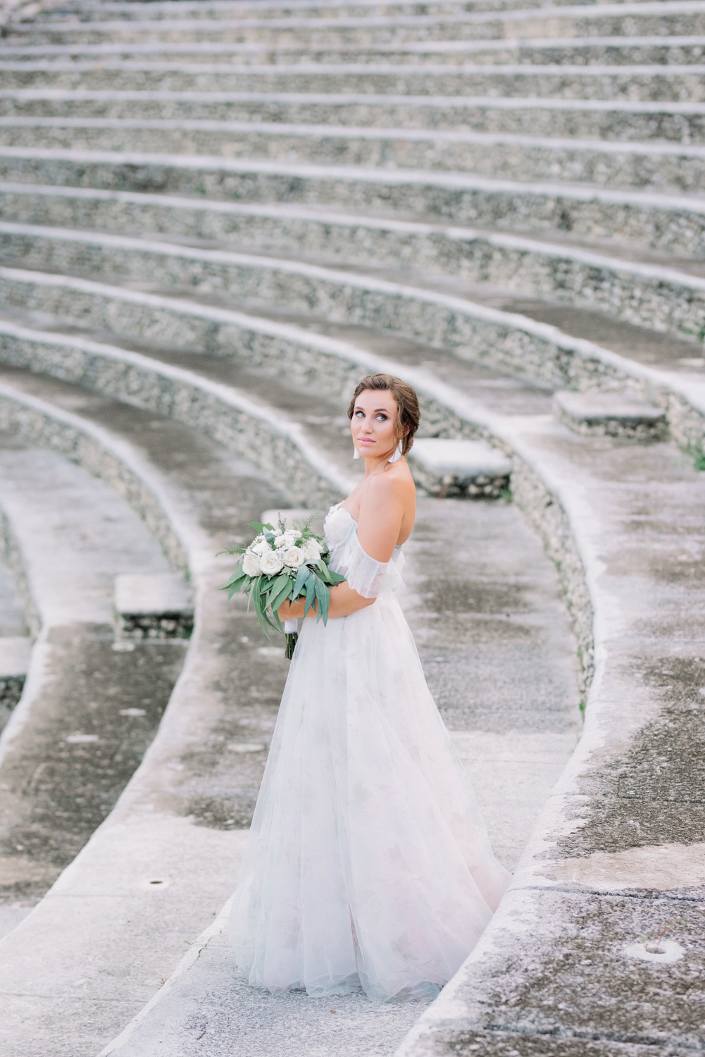 Невеста в длинном белоснежном свадебном платье на фоне каменной арочной лестницы амфитеатра.