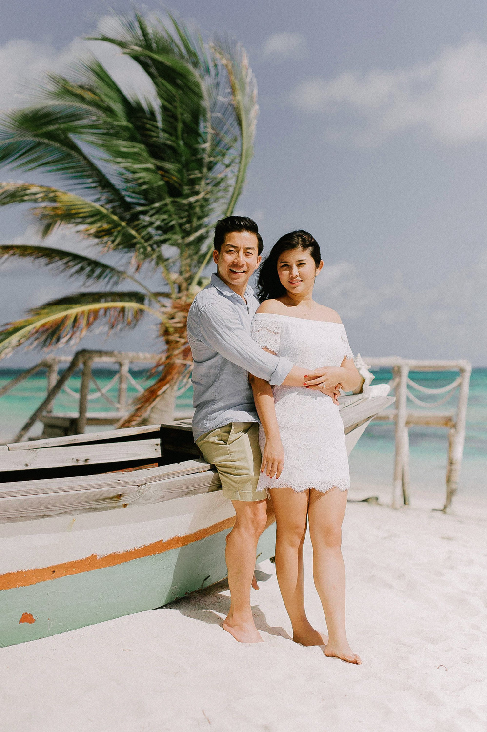 The groom hugged the bride from behind, standing by the boat under a palm tree against the background of the sea.