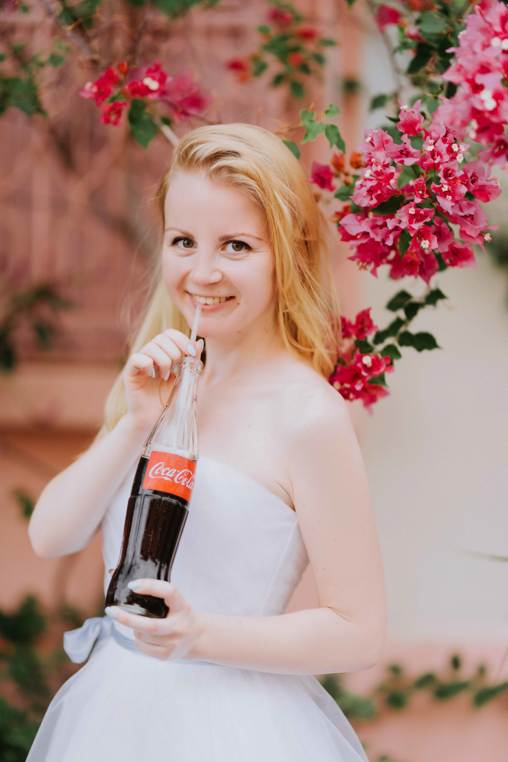 Невеста держит бутылку с напитком и трубочкой на фоне ярких красных цветов.