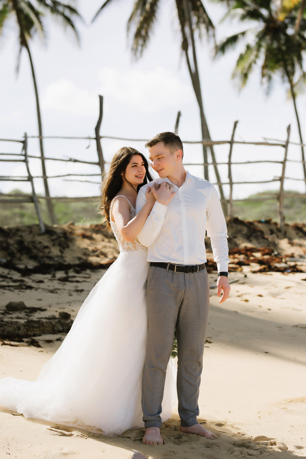 Bride and groom on the background of a fence, palm trees and blue sky.
