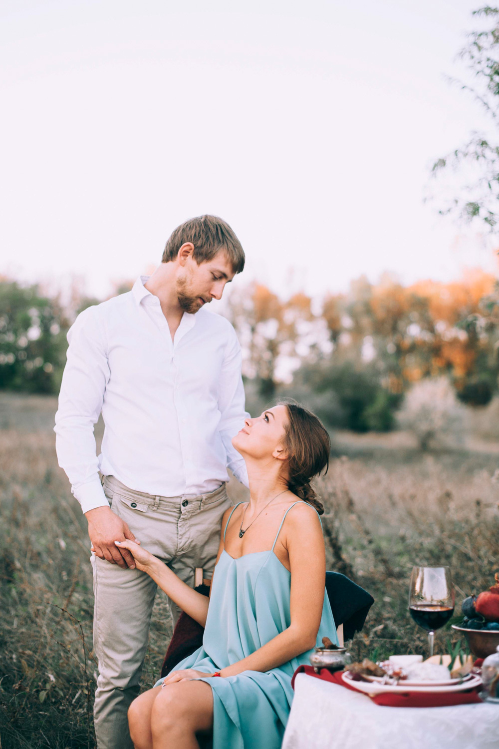 The bride sits and looks at the groom, who stands next to her and holds her hand against the background of sunlit trees.