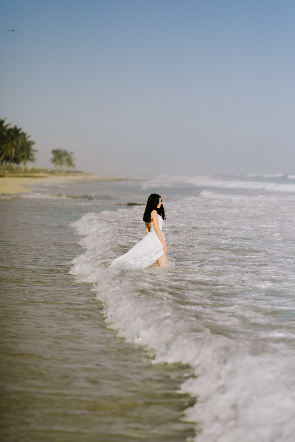 The bride looks out to sea, standing knee-deep in foaming sea water.