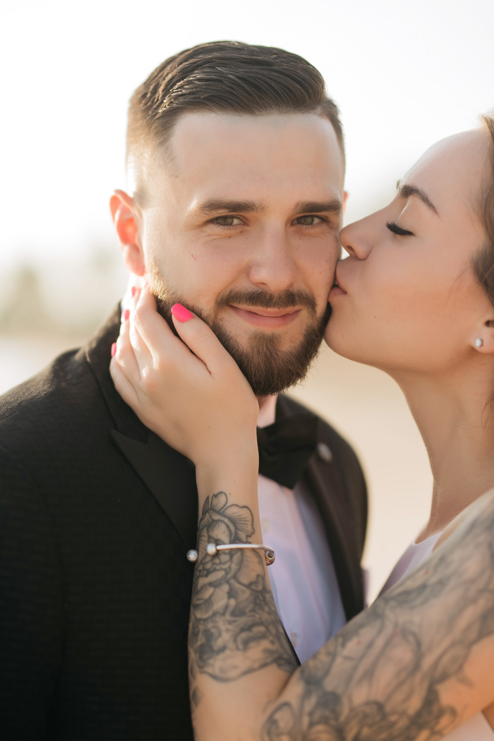 The bride embraces the groom's face with her tattooed hands and kisses him on the cheek.