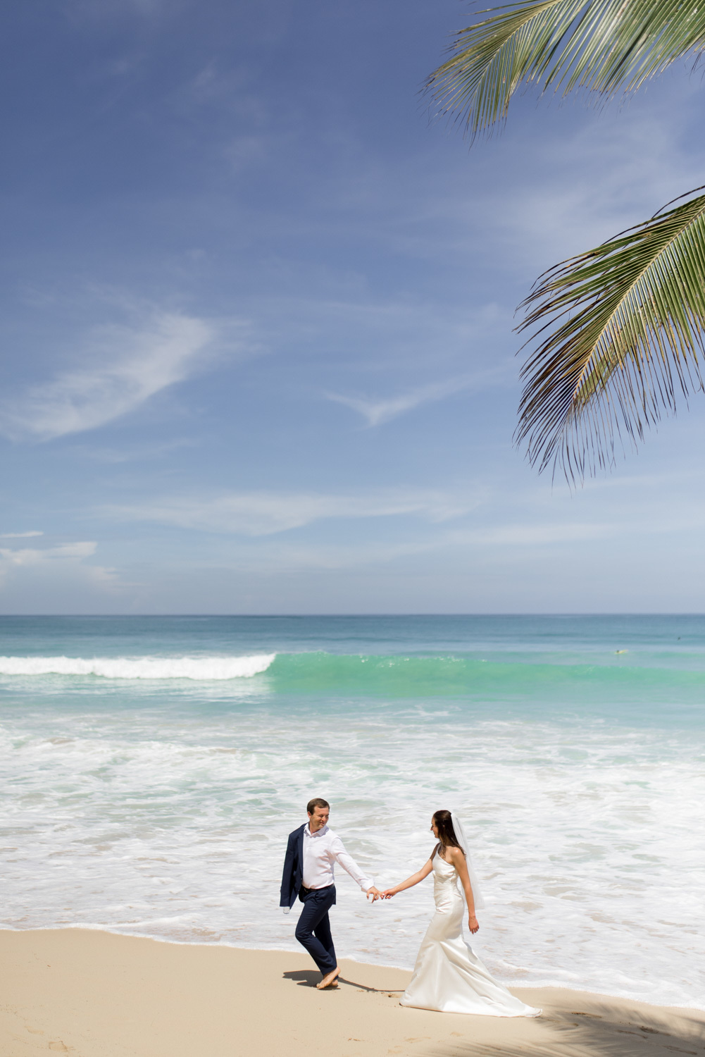 The groom leads the bride by the hand, and in the other hand he has a jacket. They walk along the sea.