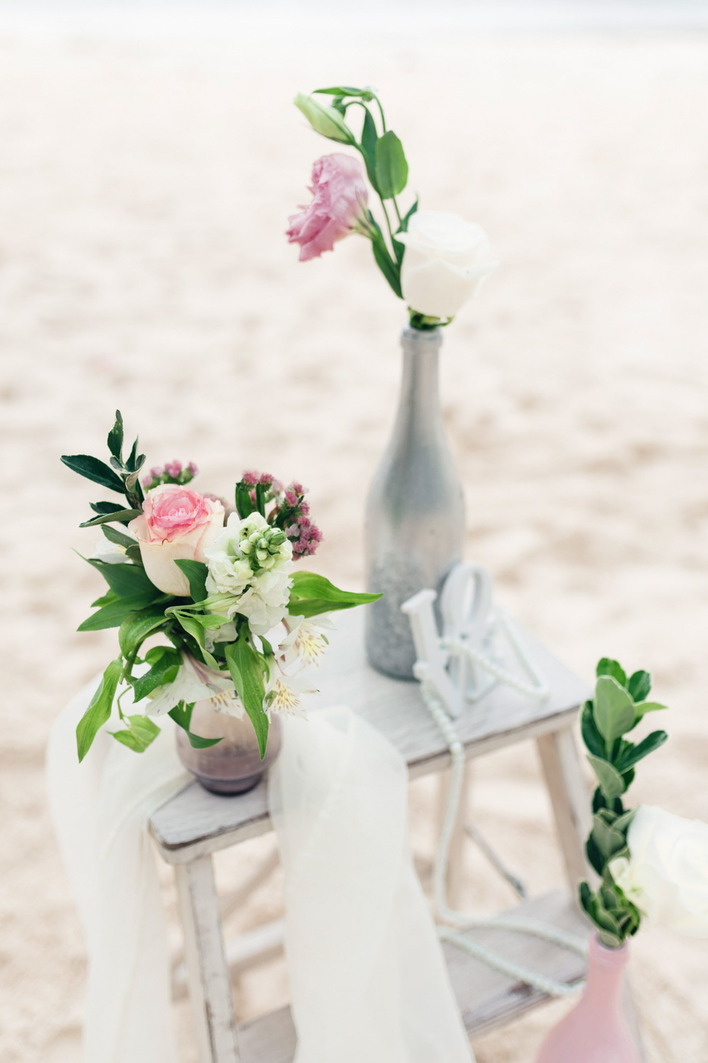 On the sand on a bench two bouquets of flowers in vases and a nylon scarf.