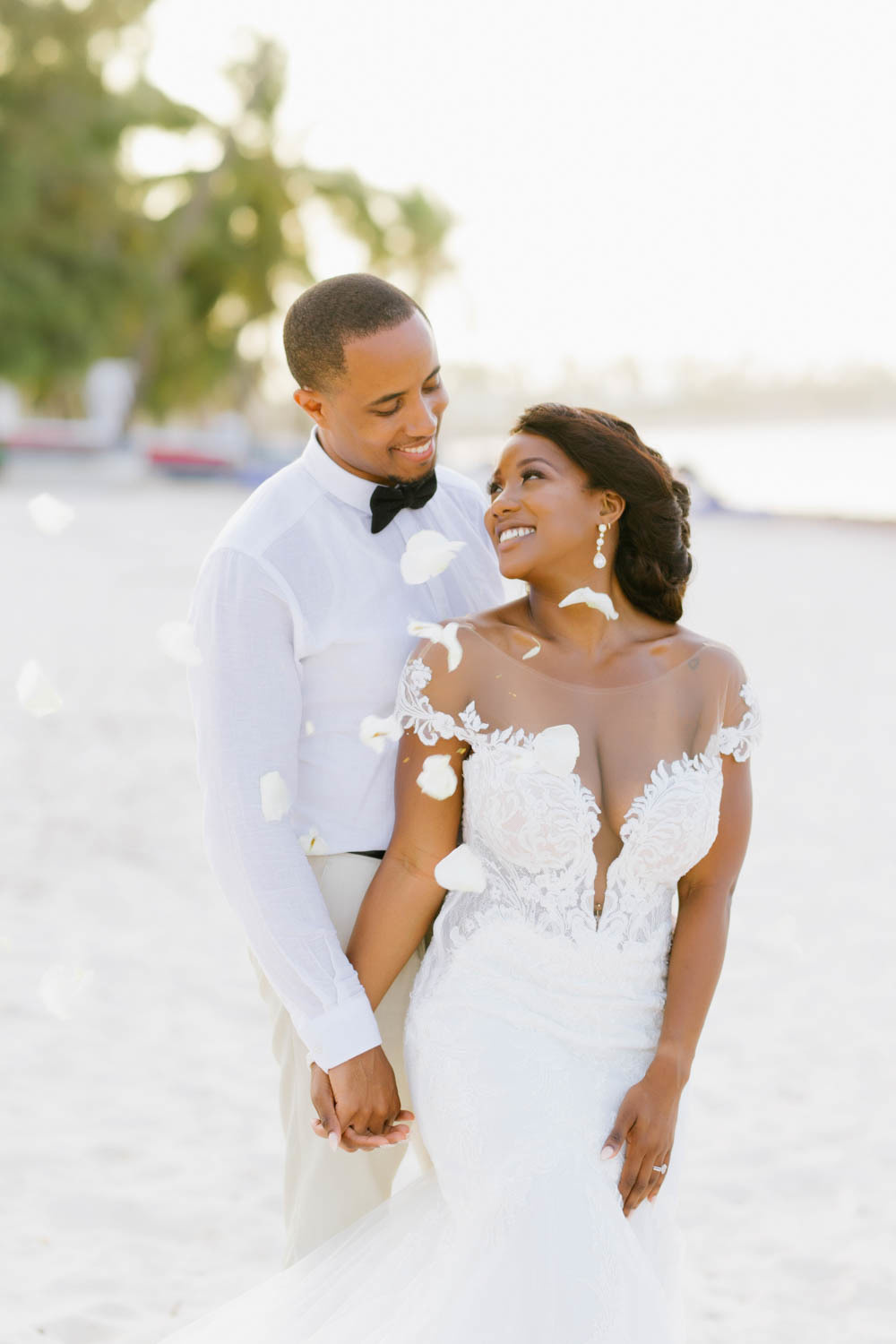 Against the background of white sand and palm trees, the bride and groom hold hands, looking into each other's eyes.