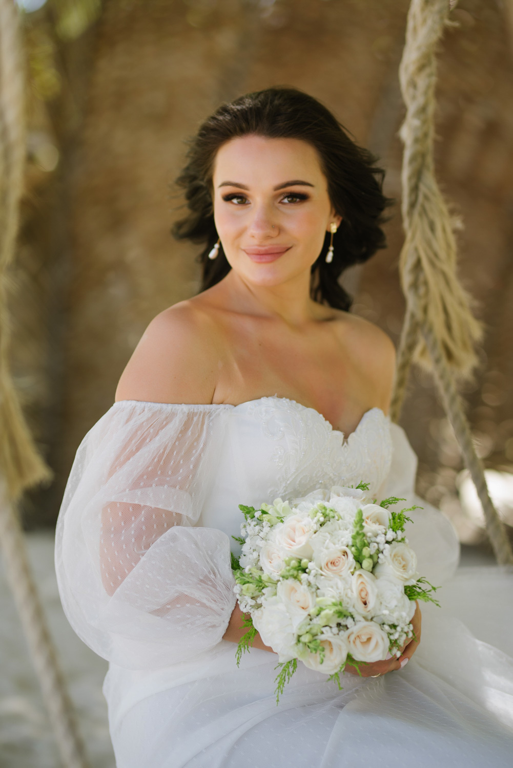 Bride in a white dress with transparent sleeves, white earrings, with a bouquet of white roses in her hands on a brown background.