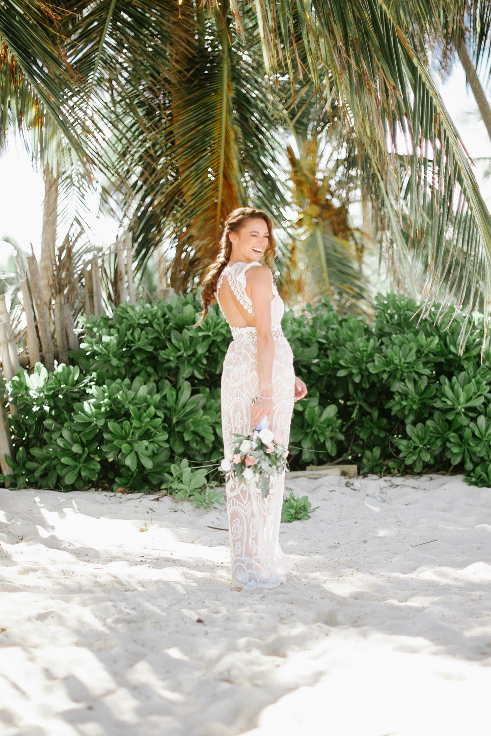 The bride with a bouquet of flowers, turning around, looks back at the background of palm trees and green shrubs.