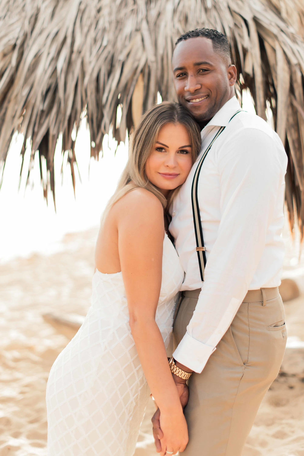 Жених с невестой, обнявшись, смотрят на фотографа на фоне пальмовой крыши пляжного домика.