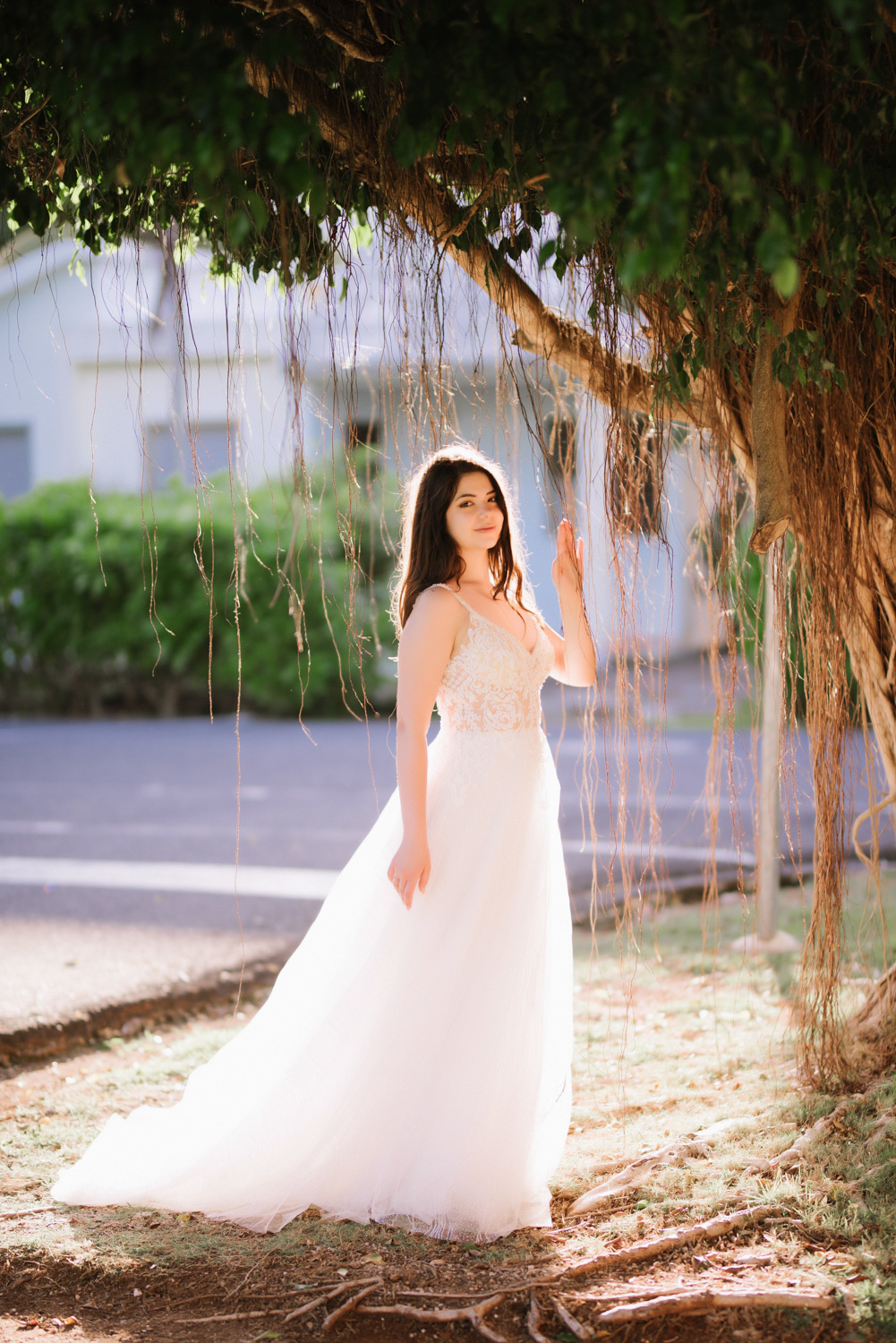 The bride stands in the shade of a tree against the background of a city building.