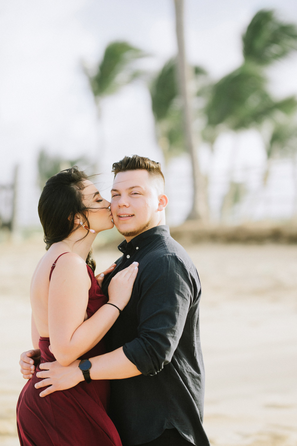 Невеста целует, обнимающего её, жениха на фоне размытого пейзажа моря и пальм.