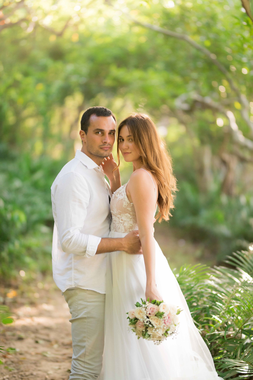 the bride and groom, who is holding a wedding bouquet, are embracing against the background of a tropical forest