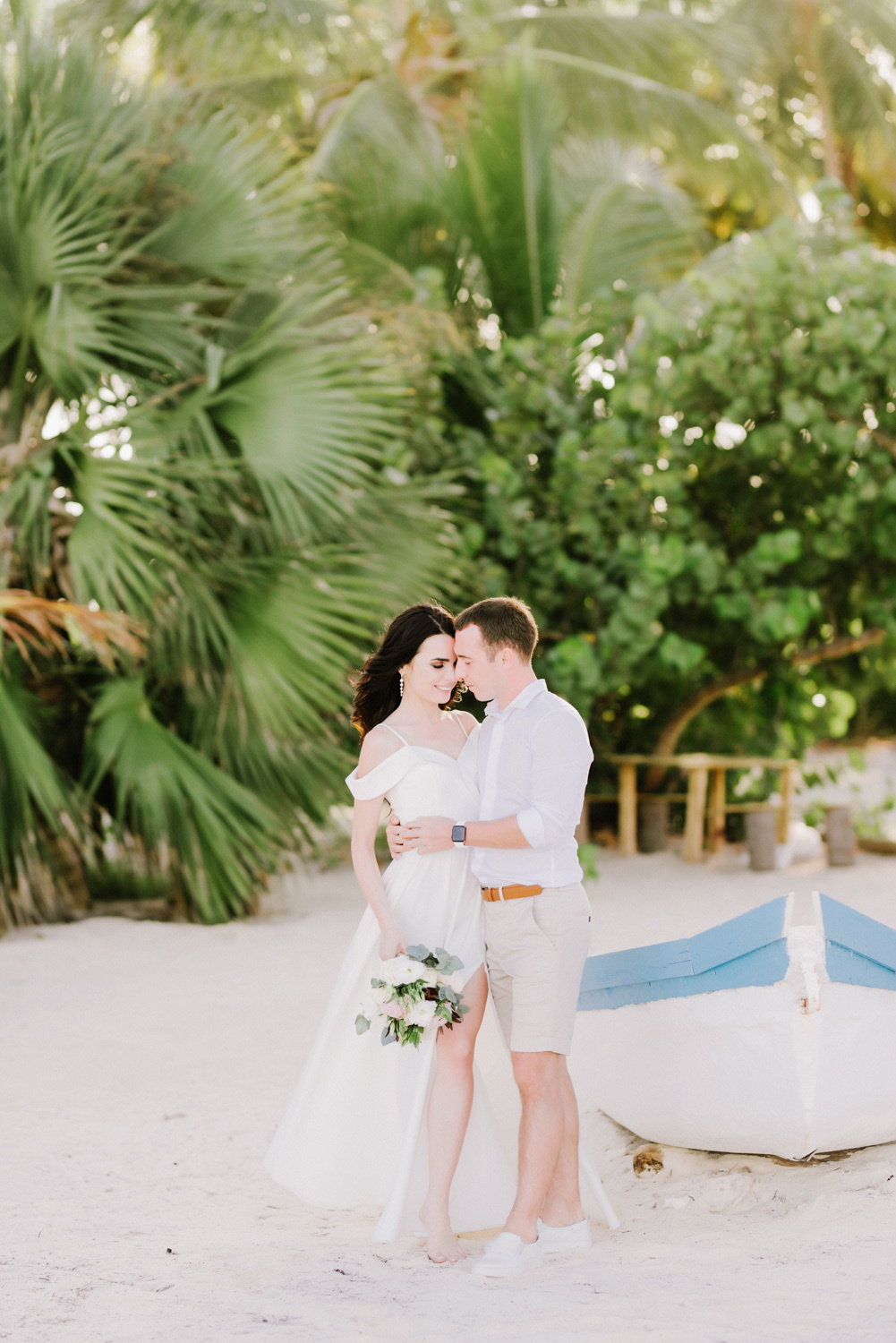 Жених обнимает невесту у лодки на фоне пальм.
