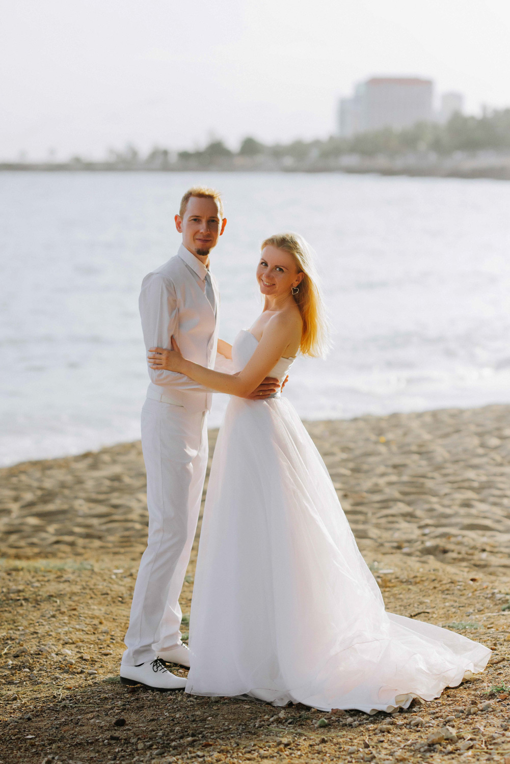 Жених с невестой держат друг друга за руки на фоне морского залива и высокого здания.
