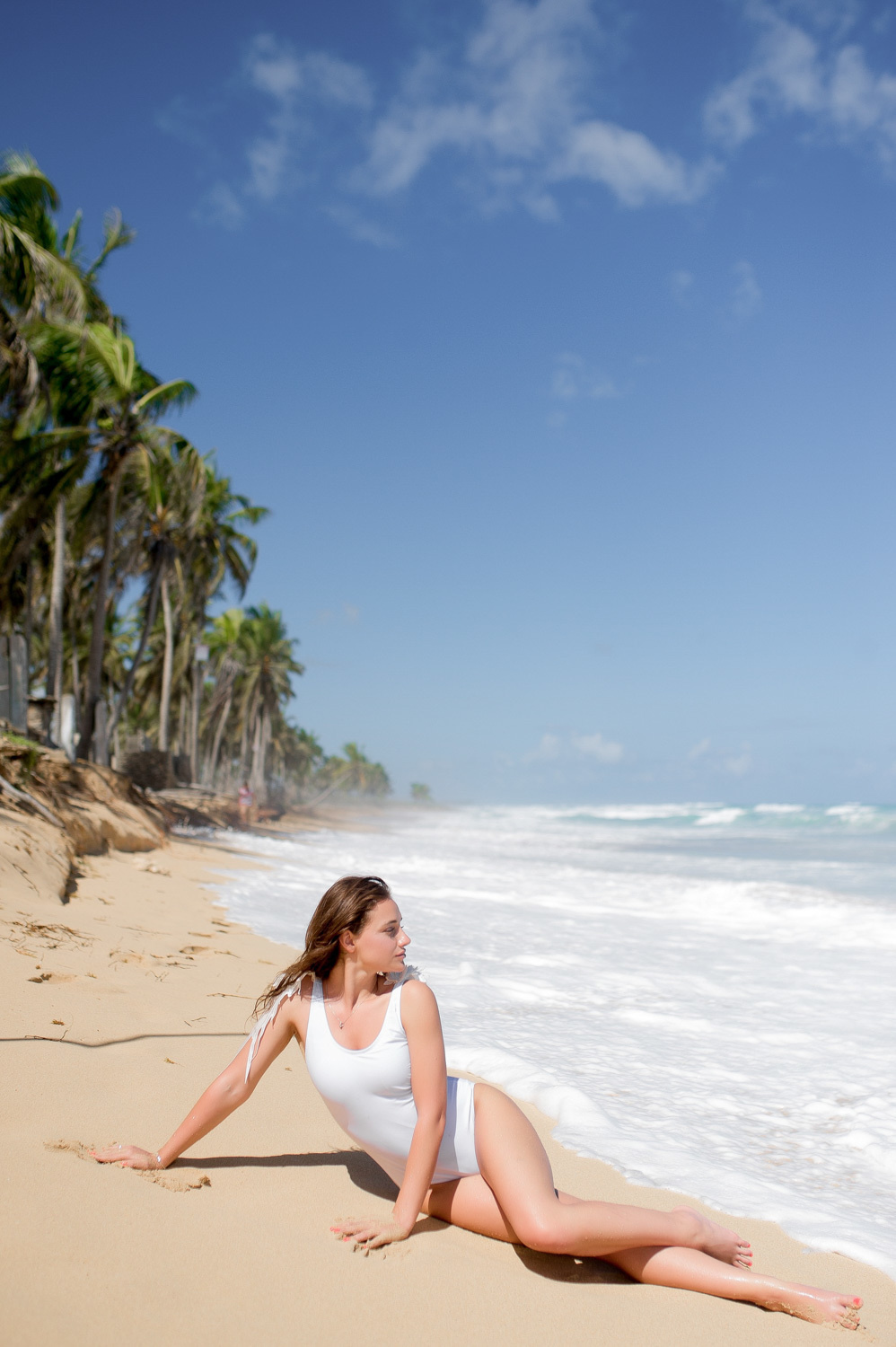 The bride in a white bathing suit lies on the sand near the sea foam against the background of coastal palm trees.