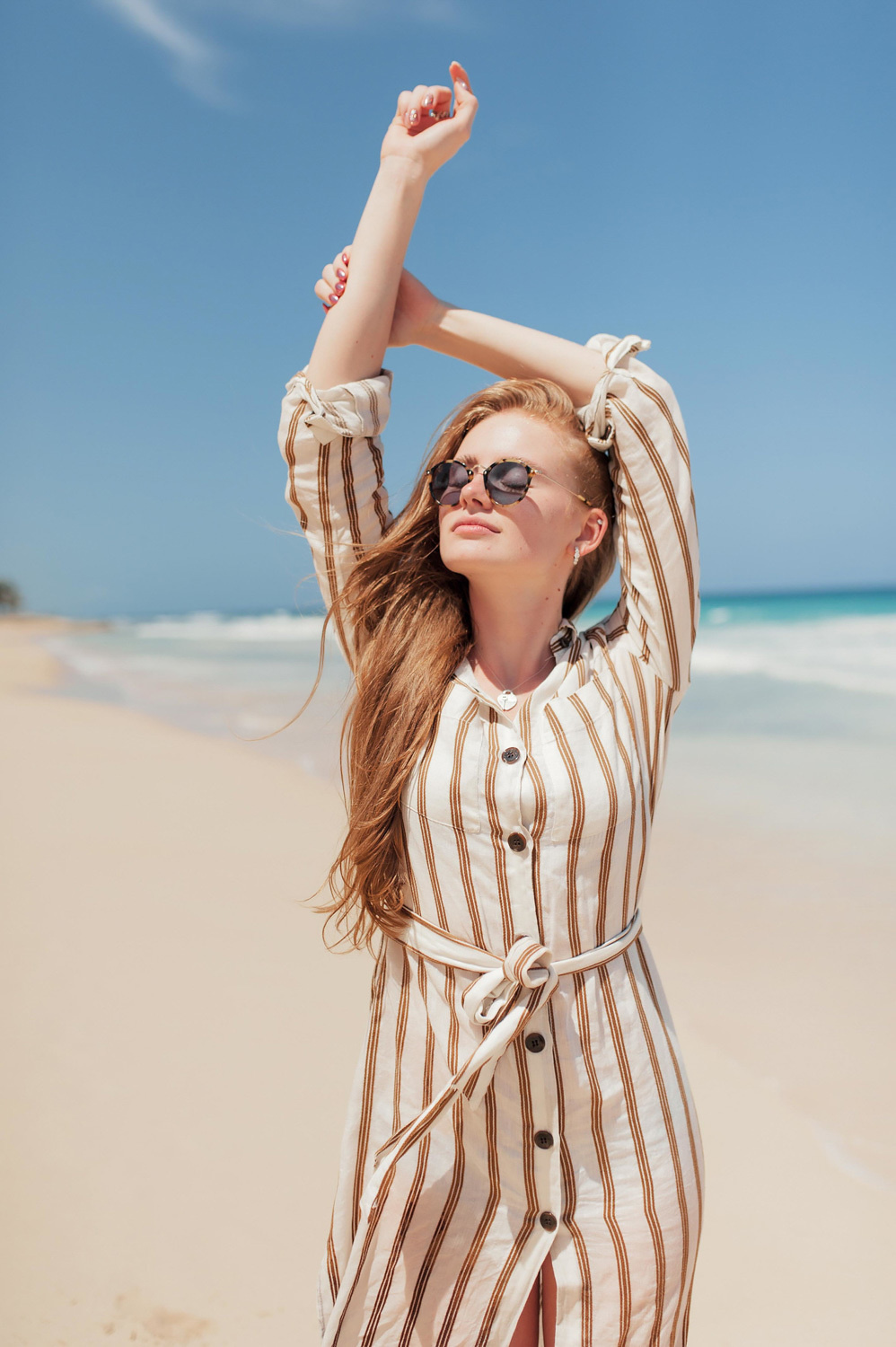 By the sea, a girl wearing sunglasses crossed her arms over her head against the sea and blue sky.