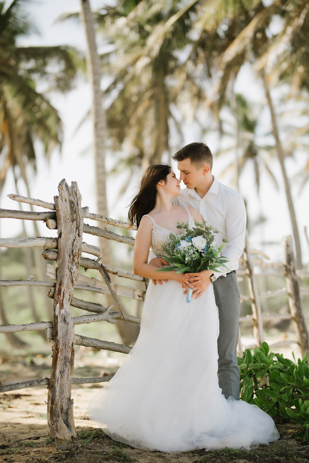 The bride in a long white dress with a bouquet in her hands is hugged by the groom at a wooden fence against the background of palm trees.