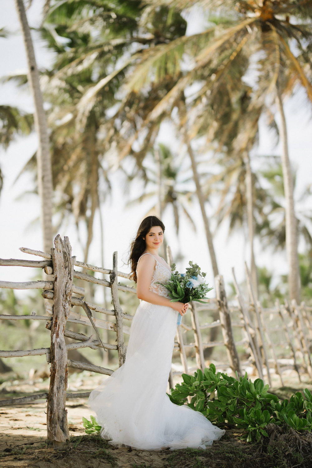 Against the background of palm trees, a bride stands at a wooden fence with a green bouquet.