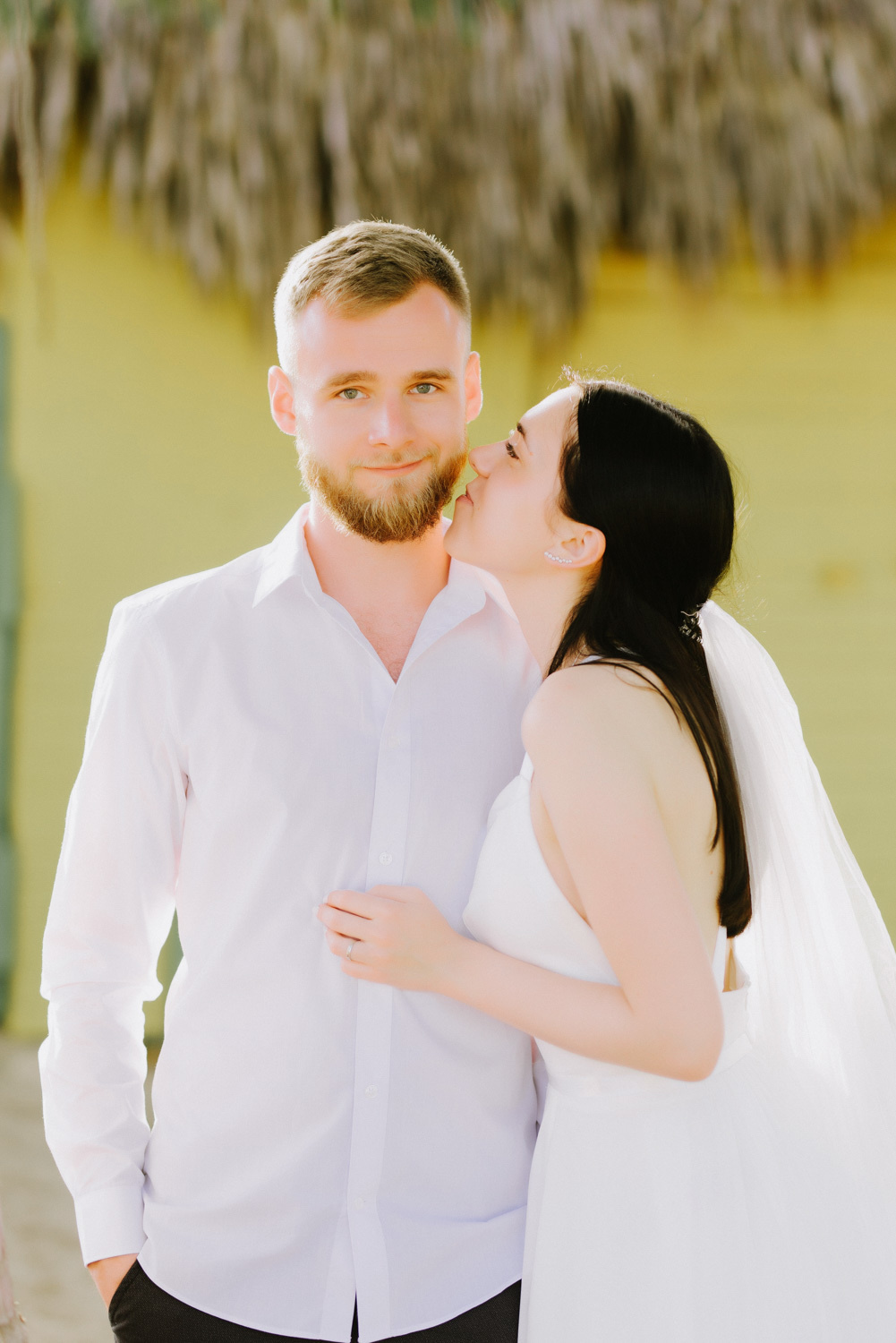 The bride kisses the groom on the cheek against the background of a yellow wall and palm leaves.
