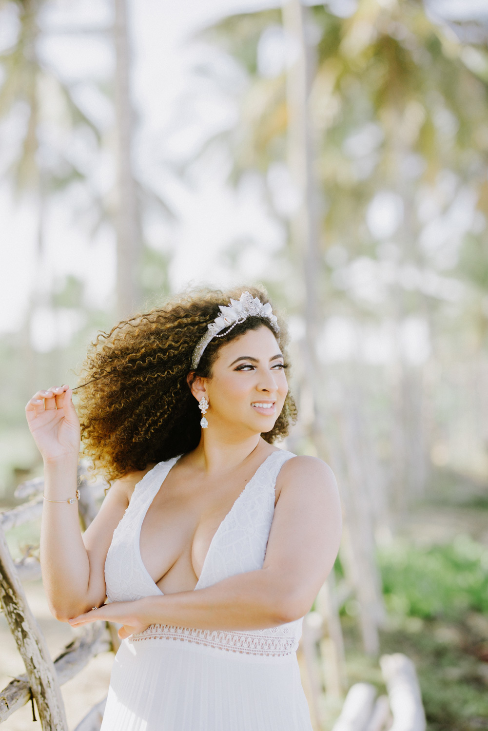 Девушка в свадебном украшении для волос на каштановых вьющихся волосах, белых длинных сережках и белом свадебном платье на фоне пальм.