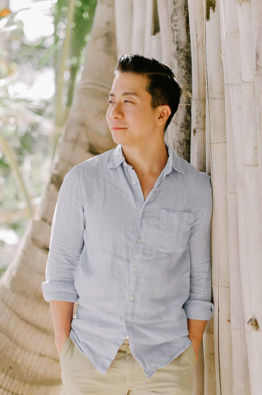 The groom holds his hands in his pockets against the background of palm trunks.