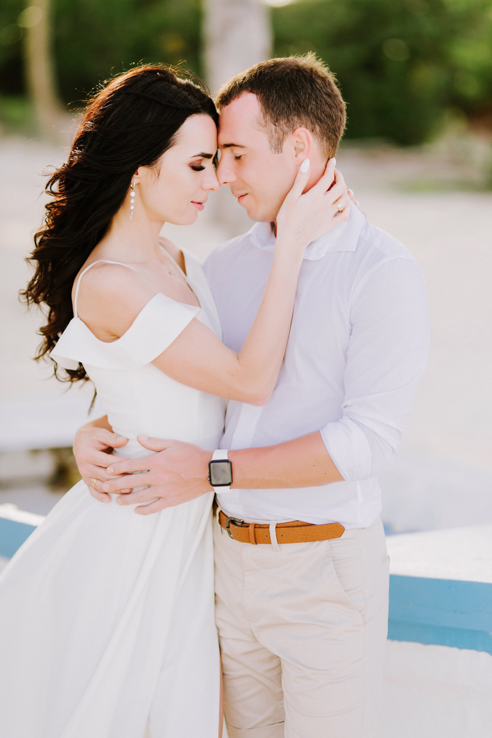 Невеста обхватила лицо жениха руками, а он обнимает её за талию на фоне пляжа.