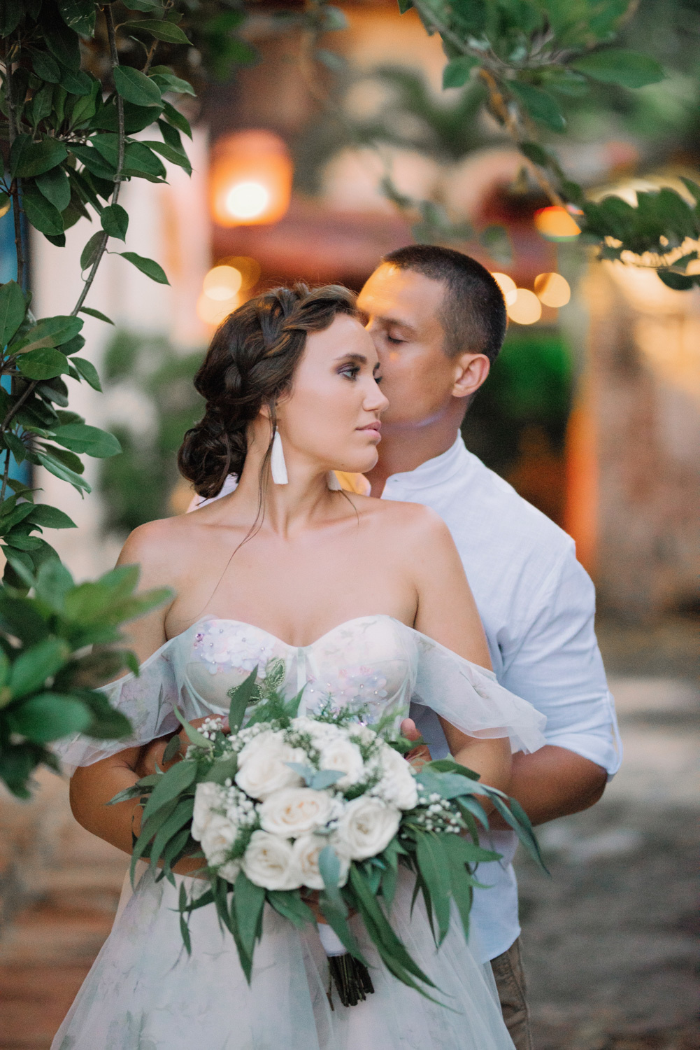 Невесту с букетом белых цветов сзади обнимает жених на фоне огней каменного храма.