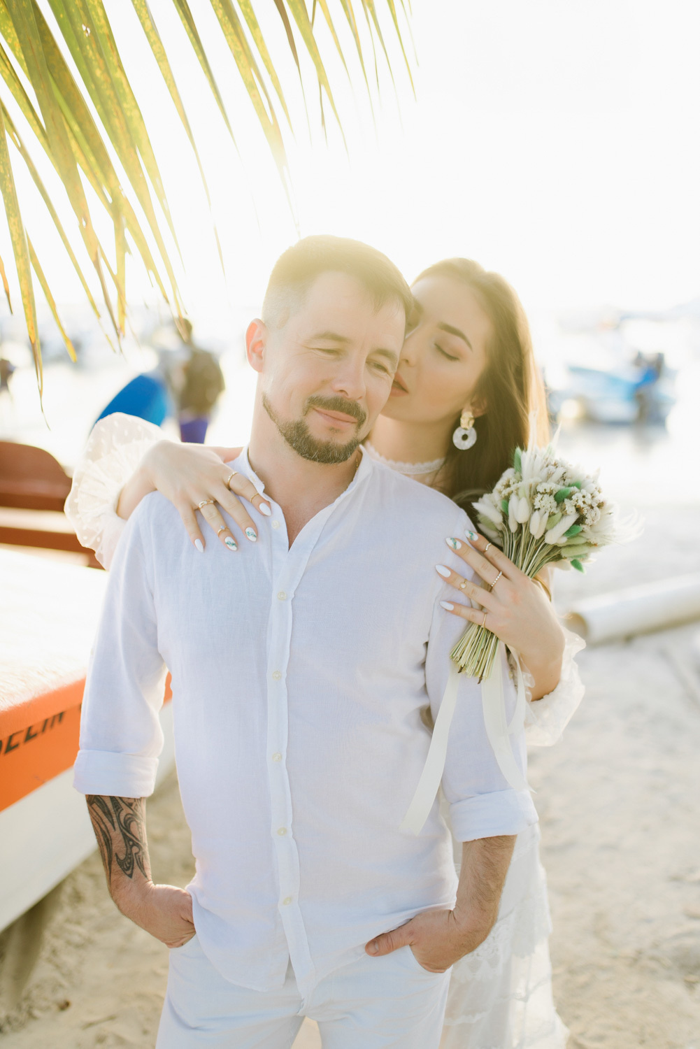 The groom with a short haircut, mustache and beard is hugged by the bride with loose hair, long white earrings in her ears, with a bouquet of white flowers on the beach background.