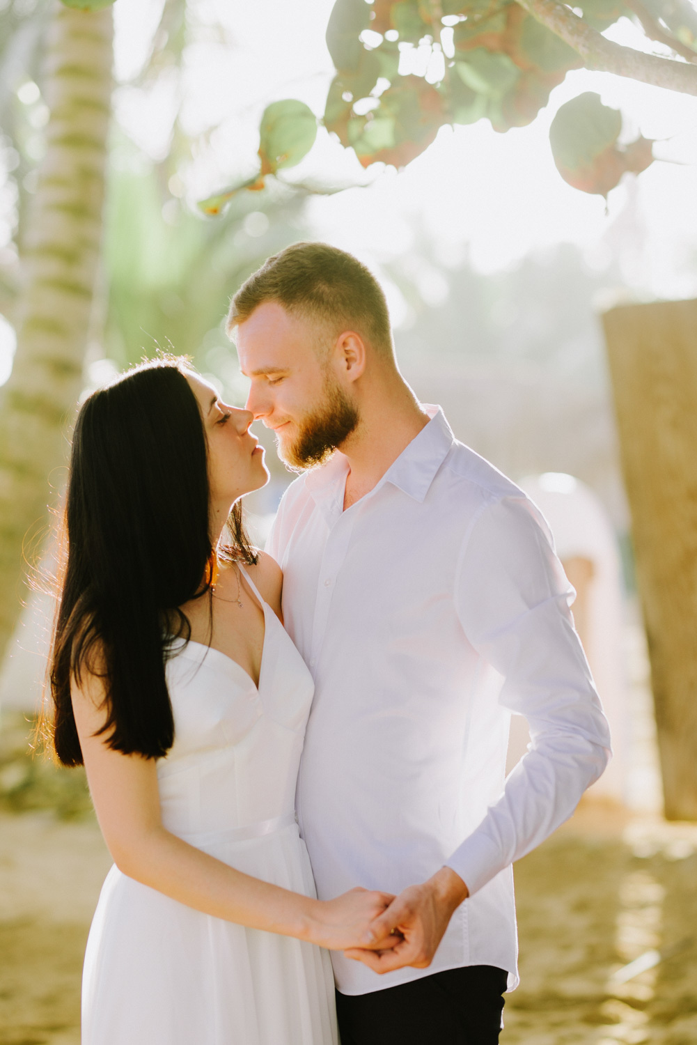 The bride with dark flowing hair and the groom with brown hair and a short haircut, with a beard hold hands and look at each other.