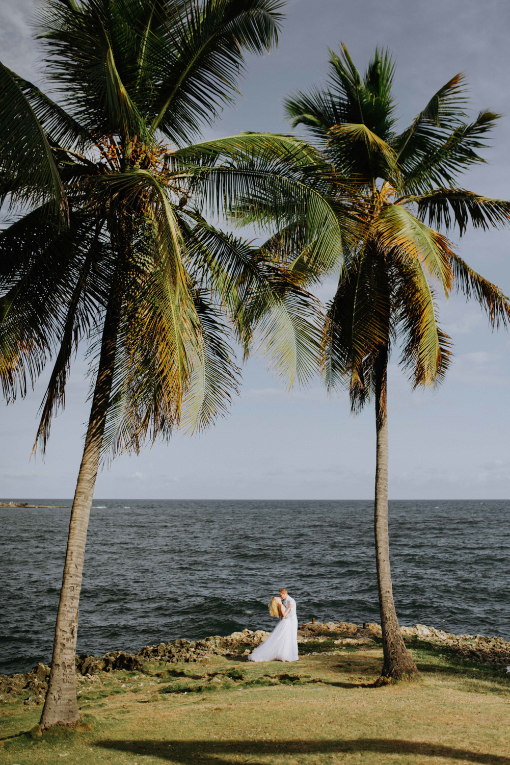 Жених с невестой стоят обнявшись между двух пальм на фоне моря.