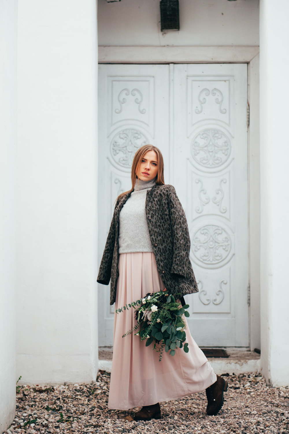 The bride is wearing a dark jacket over her wedding dress, holding a bouquet of flowers against the background of a white patterned door.