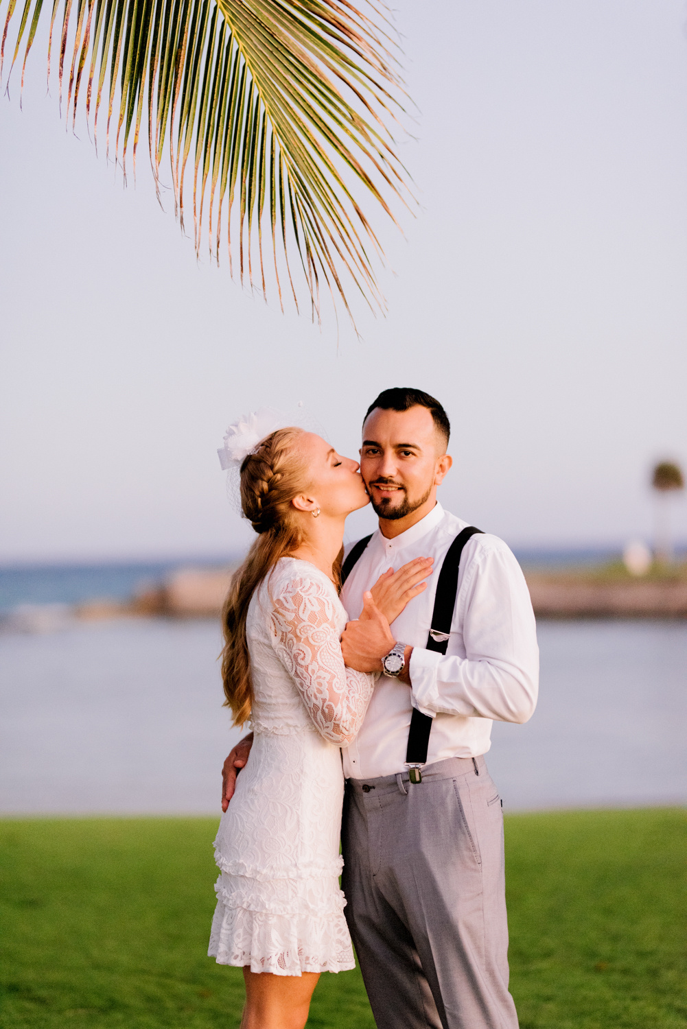 The bride, putting her hands on the groom's chest, kisses him against the background of the sea bay. Above them is a huge palm leaf.
