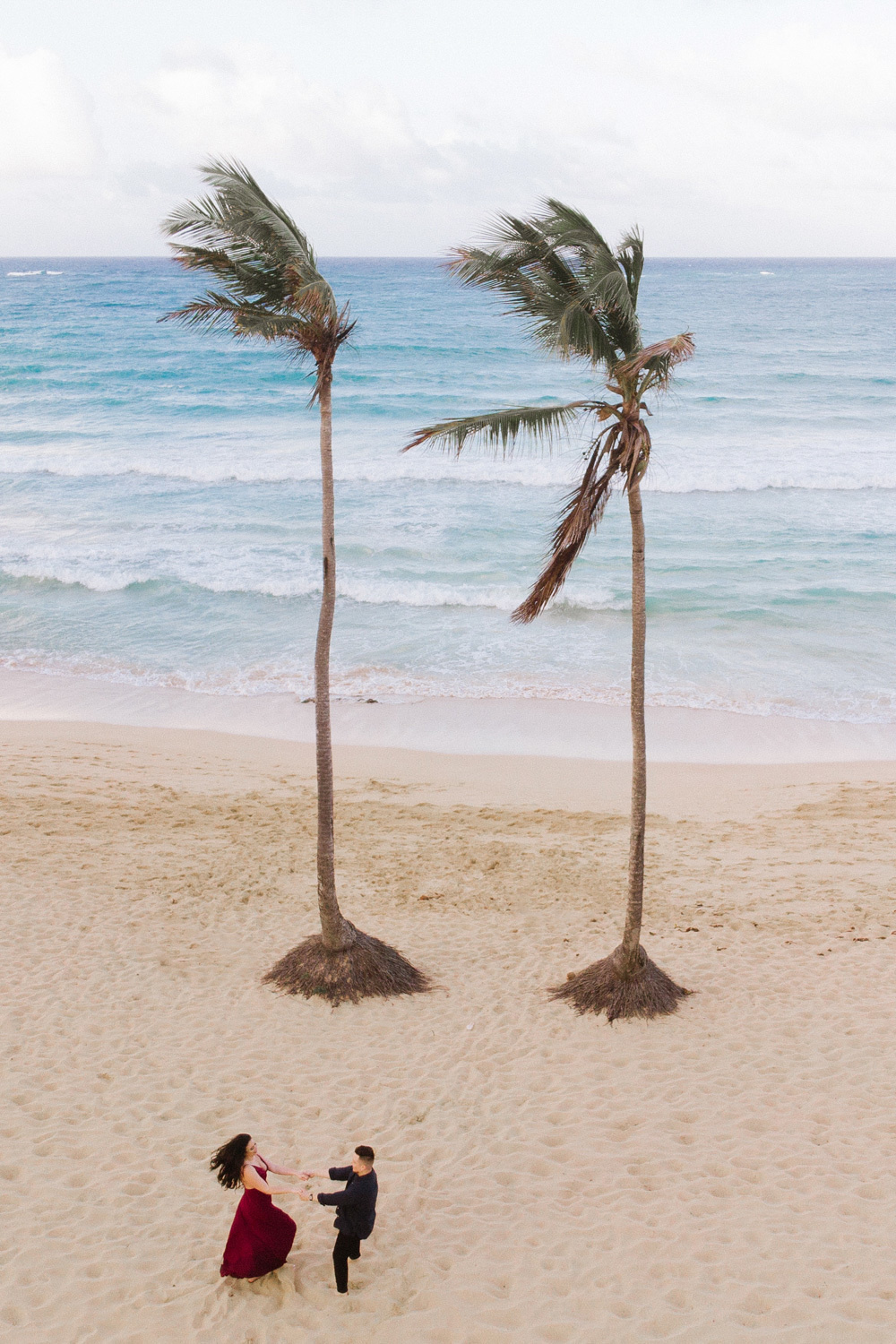 Жених с невестой держат друг друга за руки у двух пальм на фоне моря.