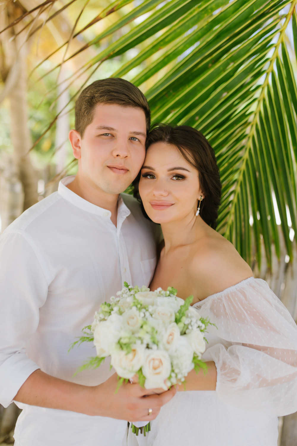 The bride and groom stand with their arms around each other, holding a bouquet of flowers in their hands, against the background of palm trees.