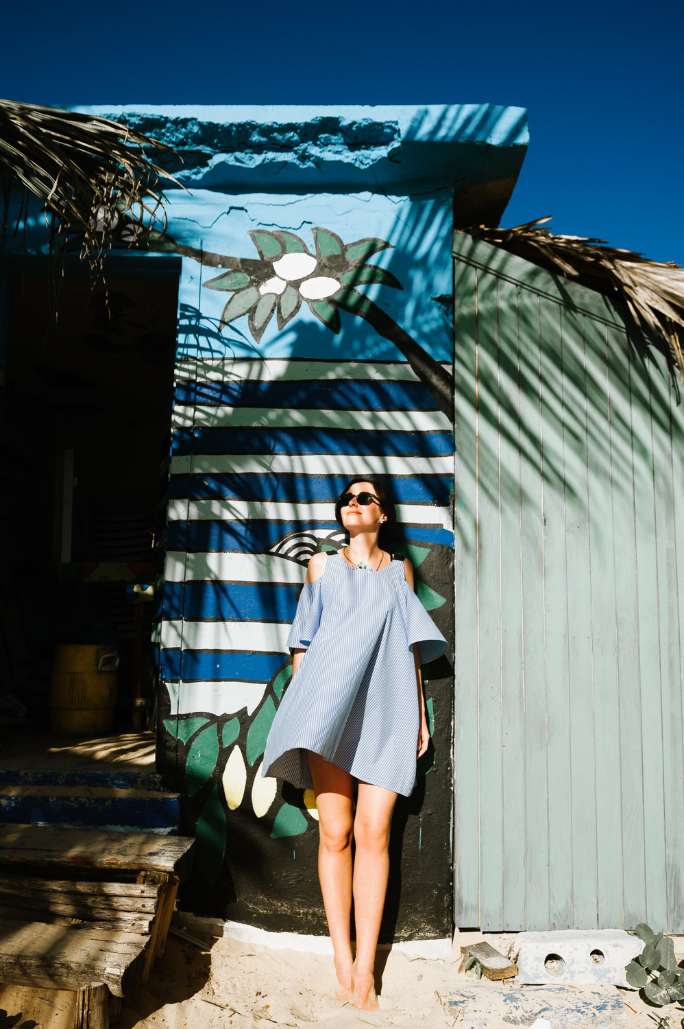 A girl in a tunic leans against the wall of a blue-painted beach house.