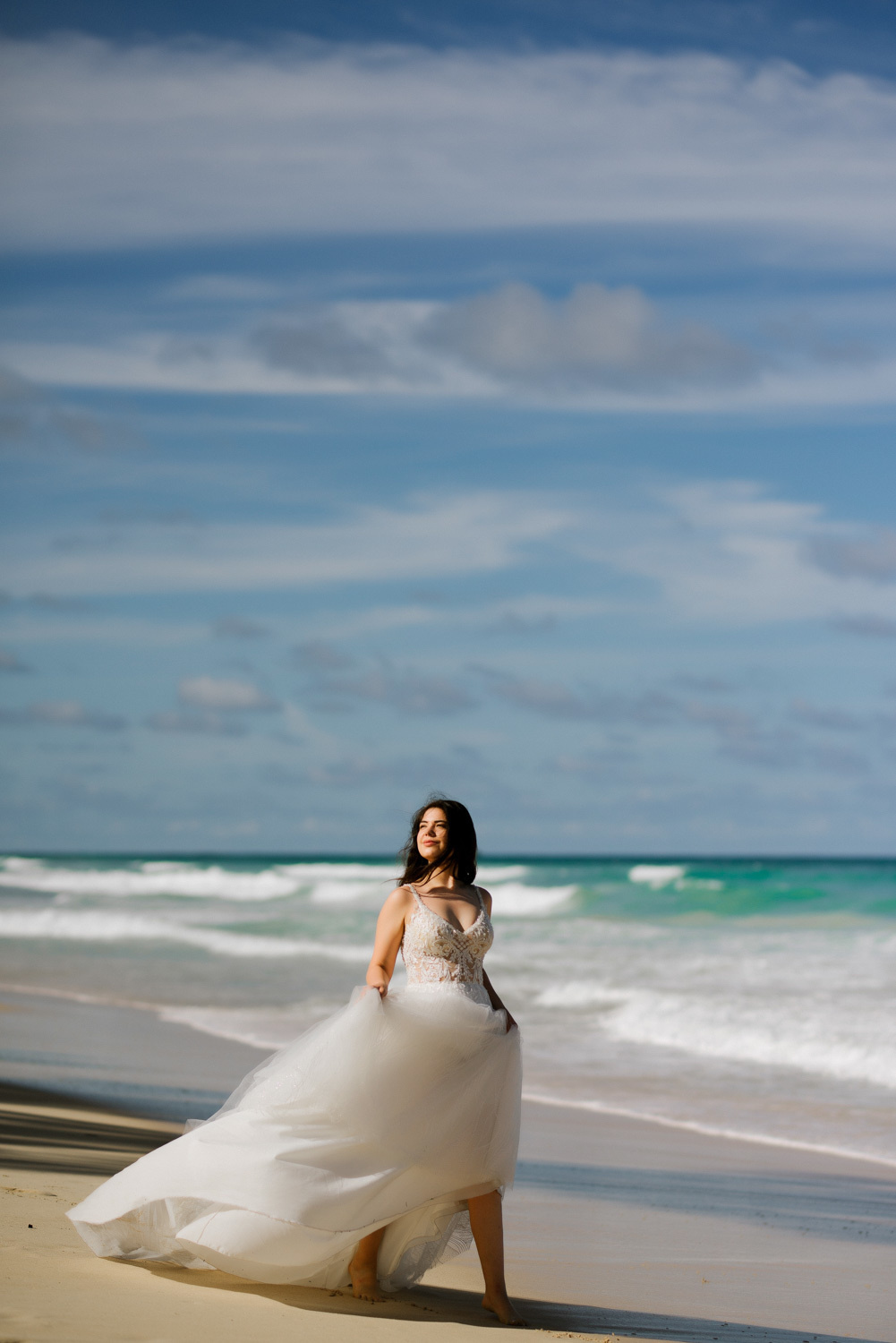 Bride in a light white dress on the background of white foam waves, turquoise sea and blue sky with white clouds.