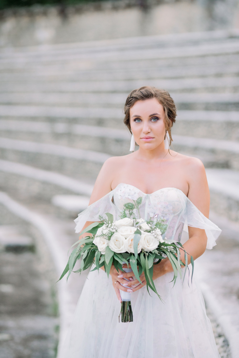 Невеста с букетом белых цветов на фоне каменной арочной лестницы.