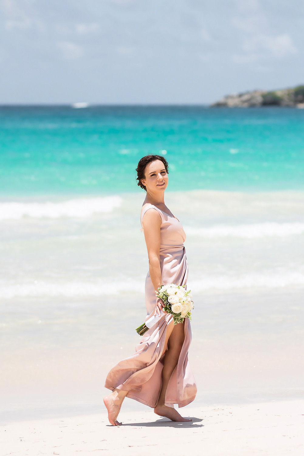 Черноволосая невеста в розовом свадебном платье с букетом цветов на пляже на фоне моря.