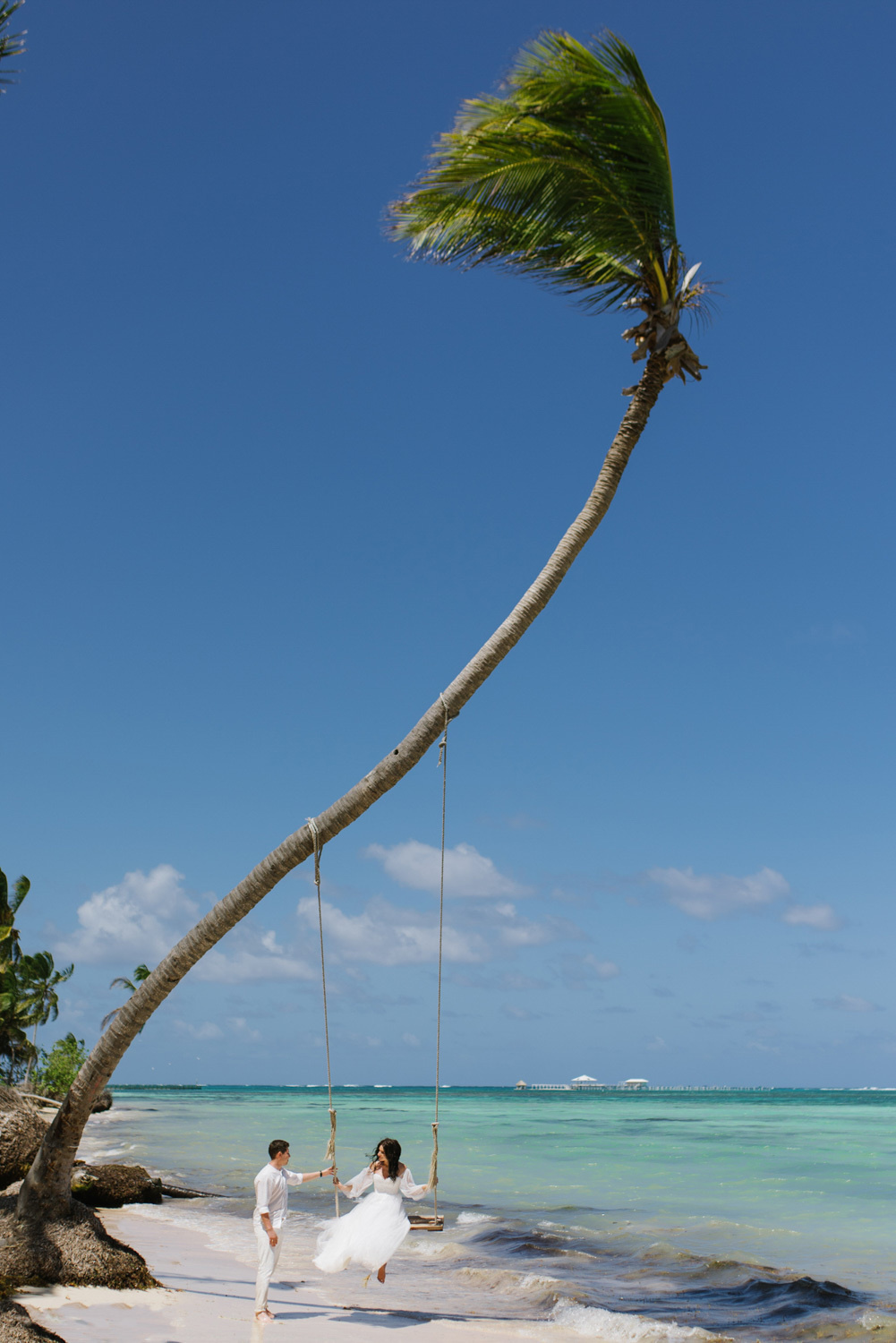 The groom swings the bride on the swing of a tall palm tree against the background of the sea.