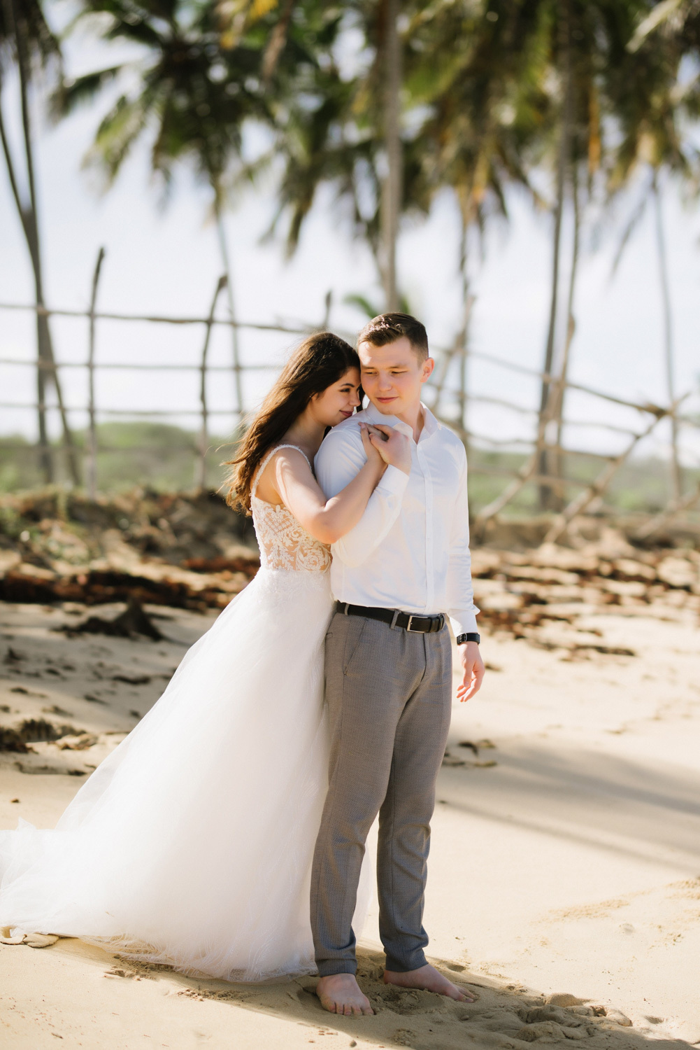 Against the background of a wooden fence and palm trees, the bride embraces the groom from behind.