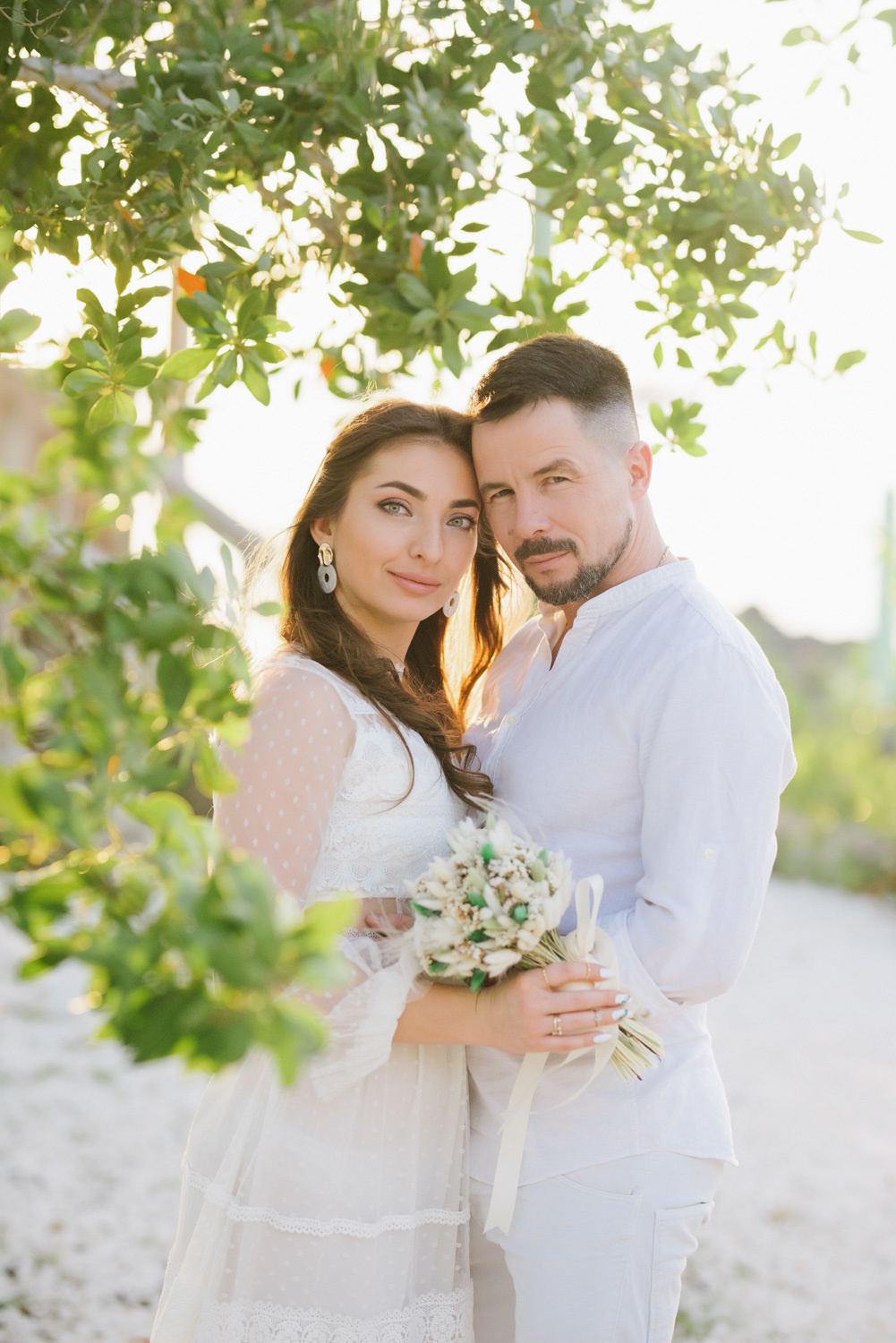 The bride with a bouquet of flowers is hugged by the groom against the background of a green tree crown.